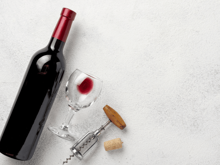 A red wine bottle lays on table next to a wine glass and a cork opener.