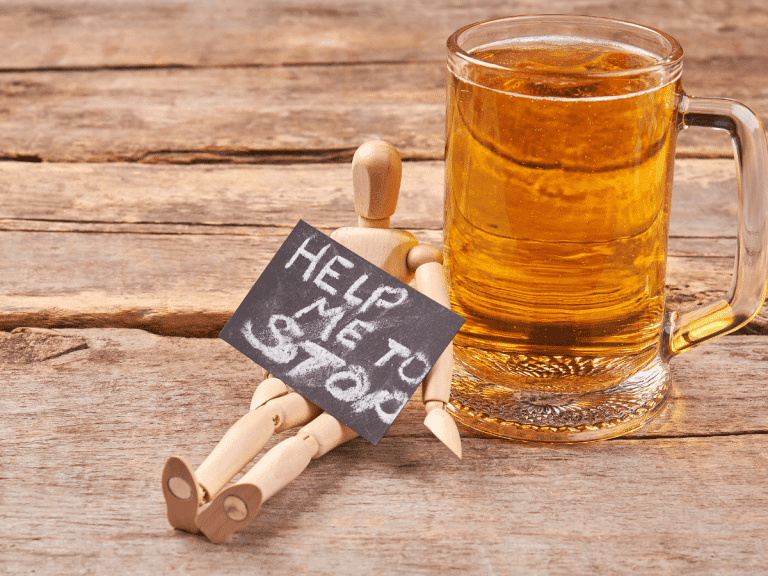 A wooden figure of a man leans against a glass of beer holding a sign that reads "help me to stop."